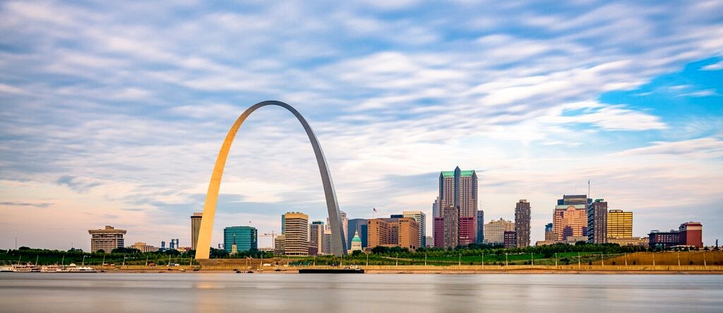A view of the St. Louis skyline and arch from across the water as the sun begins to set on a partly cloudy day.