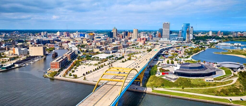 Milwaukee, WI as seen from above and across an arched bridge over the nearby river on a bright sunny day.