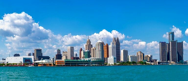 The Detroit skyline as seen from across the water on a sunny day. The sky is blue and partly cloudy.