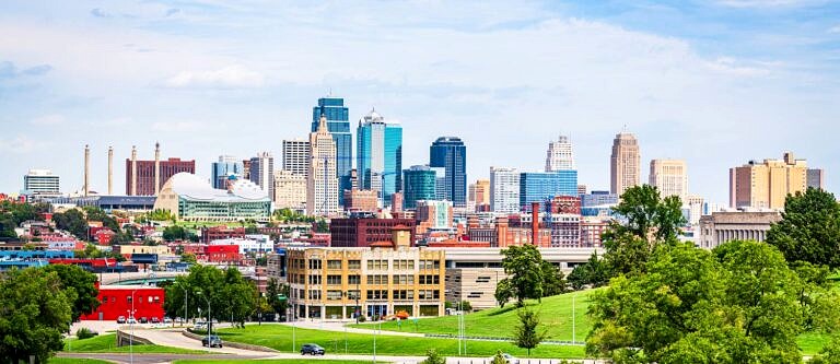 The Kansas City, MO skyline from across a grassy park and lake on a bright, partly cloudy day.