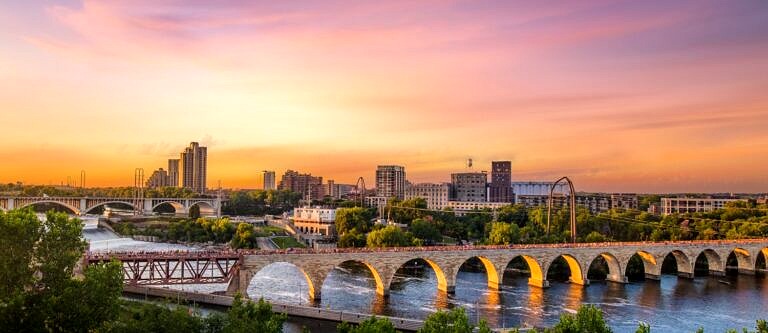 The Minneapolis skyline at dusk. The sky is orange and pink with wispy clouds and people are gathering on the brick bridge in the foreground.