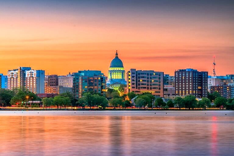 A sunset view of Madison, WI and the capital building from across the lake. The capital building is lit up with green light, and the orange sunlight coats the clouds.