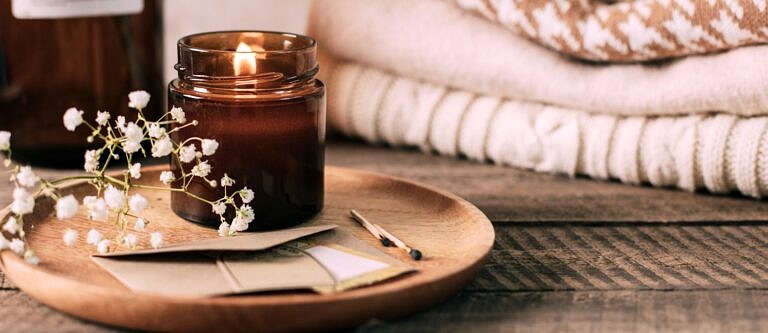 A candle in a brown glass jar sits on a small platform alongside some flowers and papers at someone's bedside.