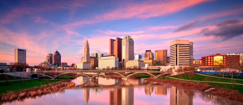 A panoramic view of the Columbus, OH skyline at sunset. The sky is blue with pink clouds streaking across, and the city is reflected across the water.